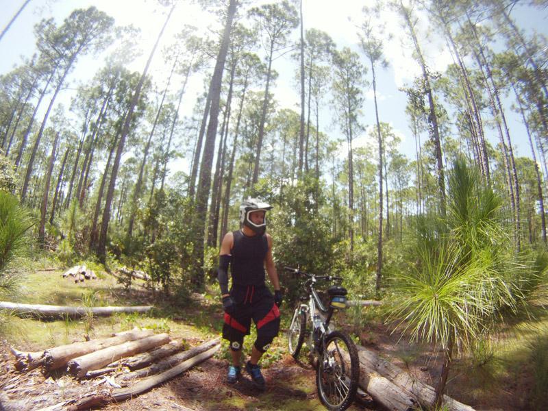 A cyclist in protective gear stands next to a mountain bike in a forested area with tall pine trees. Sunlight filters through the trees, casting dappled shadows on the ground. The cyclist wears a helmet, sleeveless shirt, and shorts, with one hand resting on the bike. Logs and greenery are visible in the foreground. The Preserve mountain bike trail.