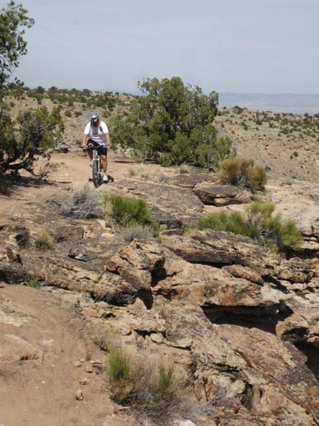A cyclist navigating a rocky trail on a hillside, surrounded by sparse vegetation and distant hills under a clear sky. Butter Knife mountain bike trail.