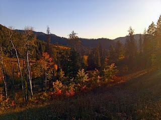 A scenic landscape featuring rolling hills and mountains in the background, surrounded by trees displaying autumn foliage. The scene is illuminated by warm sunlight, creating a tranquil and picturesque setting. Cache Creek - Game Creek Loop mountain bike trail.