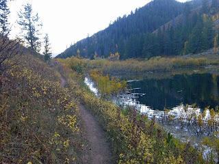 A peaceful natural landscape featuring a dirt path alongside a calm lake, surrounded by autumn foliage and evergreen trees, with mountains in the background. Cache Creek - Game Creek Loop mountain bike trail.