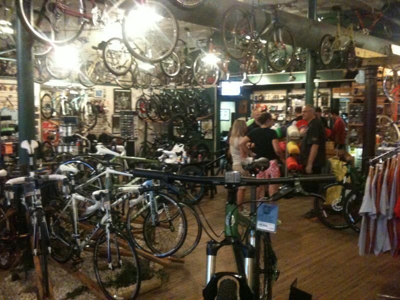 A busy bicycle shop interior with a variety of bikes displayed on walls and racks. Customers are gathered in conversation, while several bicycles, including road and mountain bikes, are positioned throughout the space. The shop features wooden flooring and bright overhead lighting, creating a lively shopping atmosphere.
