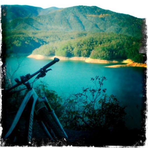 A mountain bike resting on a rocky ledge, overlooking a serene lake surrounded by lush green hills and mountains in the background. The scene captures a peaceful outdoor setting, evoking a sense of adventure and tranquility. Tsali Right Loop mountain bike trail.