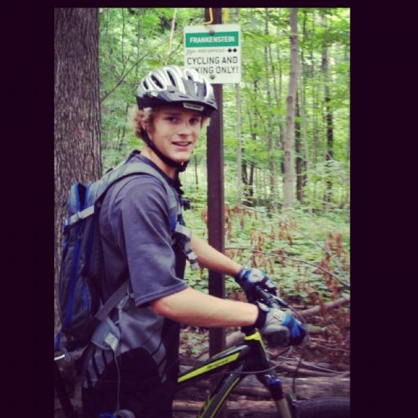 A young person with curly hair wearing a helmet and a gray shirt stands next to a mountain bike in a forested area. In the background, there is a sign indicating "FRANKENSTEIN - CYCLING AND HIKING ONLY." The setting features lush green trees typical of a biking or hiking trail. Hydrocut mountain bike trail.
