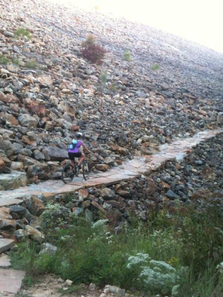 A mountain biker rides along a narrow stone path that cuts through a rocky landscape. The environment features a steep incline covered in various sizes of stones, while patches of greenery and wildflowers are visible at the base of the slope. Raccoon Mountain Trail Network mountain bike trail.