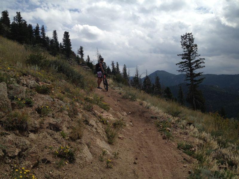 A mountain biker standing beside their bike on a dirt trail, surrounded by grassy hills and pine trees. The landscape features rolling mountains in the background beneath a cloudy sky. Walker Ranch mountain bike trail.