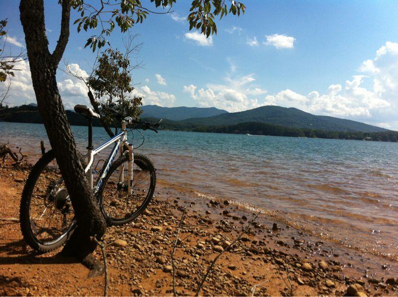 A mountain bike leaning against a tree at the edge of a serene lake, with clear blue water and rolling hills in the background under a cloudy sky. The shoreline is rocky and sandy, creating a peaceful outdoor scene. Jack Rabbit Trails mountain bike trail.