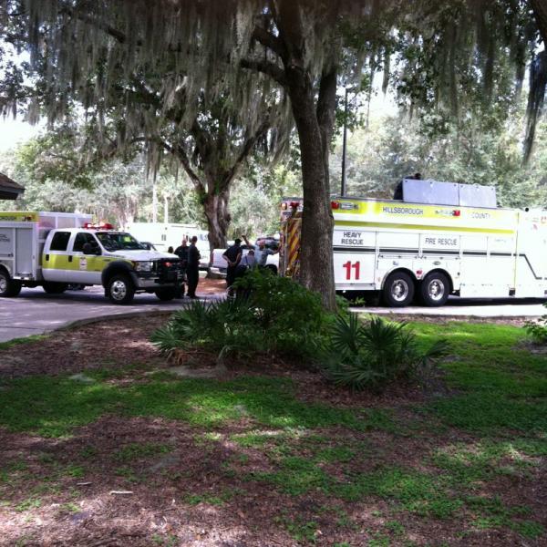 A fire rescue truck and an emergency vehicle are parked in a shaded area surrounded by trees. Emergency responders are gathered near the vehicles, discussing a situation. The scene is set in a park-like environment with lush greenery and Spanish moss hanging from the trees. Alafia River State Park mountain bike trail.