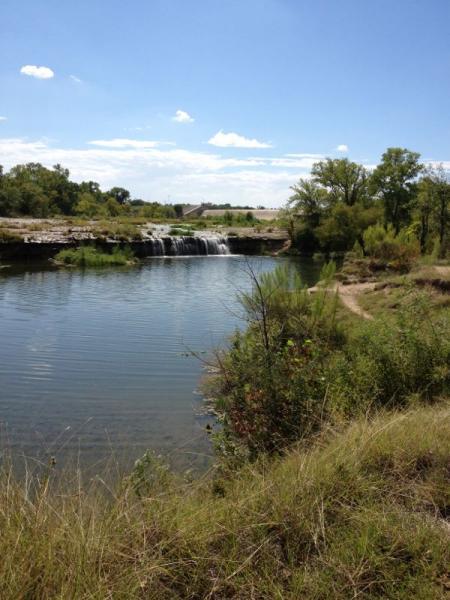 A serene landscape featuring a calm river with a gentle waterfall on the left. Surrounding the water are lush green trees and grasses under a clear blue sky with a few scattered clouds. The scene captures the beauty of nature in a tranquil setting. Sansom Park mountain bike trail.
