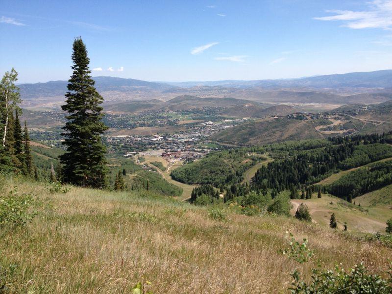 A panoramic view of a mountainous landscape featuring rolling hills and a small town nestled in the valley. Tall evergreen trees are in the foreground, with lush greenery and open grassy areas. The sky is clear with few clouds, and distant mountains can be seen beyond the valley. Mid Mountain mountain bike trail.