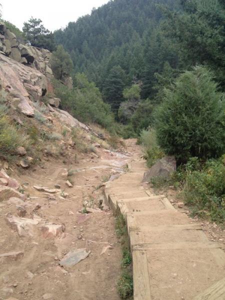 A dirt hiking path winding through a forested area, featuring wooden steps descending along the trail. The surrounding landscape includes rocky outcrops and dense greenery, with mountains visible in the background under an overcast sky. Deer Creek Canyon mountain bike trail.