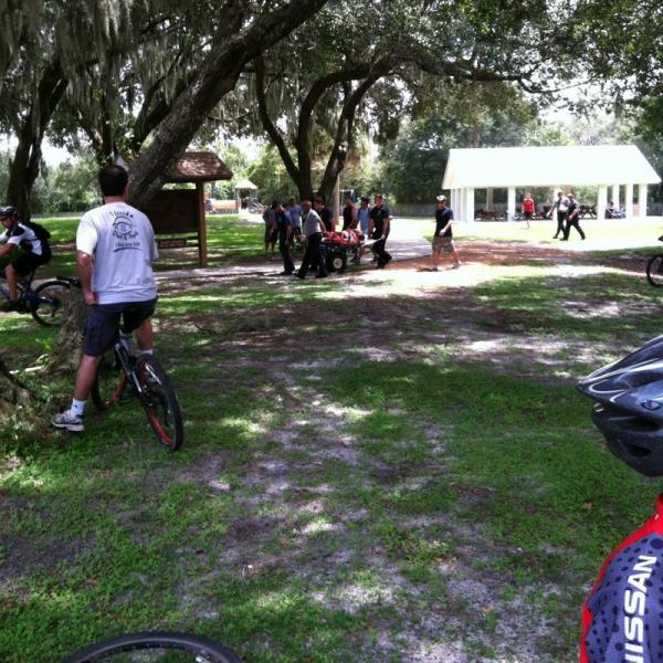 A group of people gathered in a park setting, with some standing near a bike, while others are engaging around a cart filled with items. Trees provide shade in the background, and a covered pavilion is visible in the distance. The ground is grassy, and the atmosphere appears lively and casual. Alafia River State Park mountain bike trail.