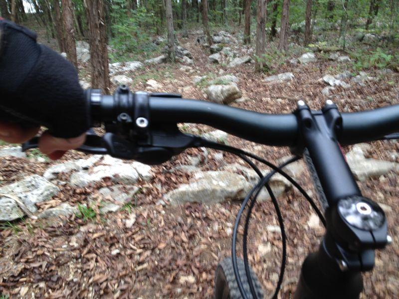 Close-up view of a mountain bike's handlebars with one hand gripping the brake, set against a rocky, wooded trail. The ground is covered with fallen leaves, and trees are visible in the background. Hamilton Creek mountain bike trail.