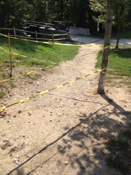 A dirt path leading through a green area is cordoned off with yellow caution tape. In the background, partially obscured by trees, several wooden structures or benches are visible, along with a faint outline of a paved walkway. Mines Falls Park mountain bike trail.