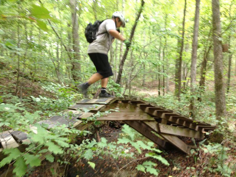 A person wearing a helmet and gloves is navigating a wooden bridge in a forested area, surrounded by lush greenery and trees. The individual is mid-step, demonstrating balance while traversing the bridge. Montgomery Bell State Park Mtb Trail mountain bike trail.