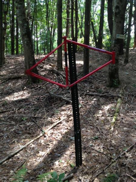 Red geometric frame mounted on a black post, set in a wooded area with trees and leaf litter surrounding it. Lippman Park mountain bike trail.