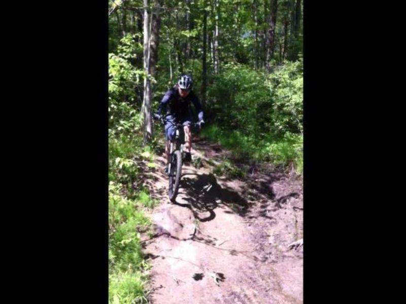 A person riding a mountain bike on a rugged, muddy trail surrounded by lush green foliage and trees. The cyclist is wearing a helmet and is navigating over rocky terrain, showcasing an action shot of outdoor biking. Cemetery Loop mountain bike trail.