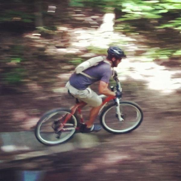 A person riding a mountain bike along a narrow trail in a forest, captured in motion to convey speed. The cyclist is wearing a helmet and a backpack, navigating over a wooden plank while surrounded by lush greenery. Frick Park mountain bike trail.