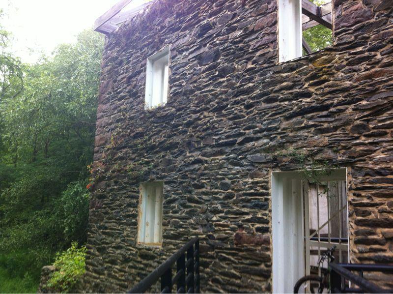 A weathered stone building with partially open windows, surrounded by lush greenery. The image shows the textured stone wall and hints of vegetation climbing up the side, suggesting a natural and rustic environment. Seneca Creek State Park Trail mountain bike trail.