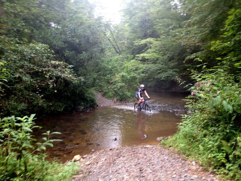 A mountain biker crosses a shallow stream surrounded by dense greenery and trees, with mist in the air creating a serene outdoor atmosphere. Jake Mountain Trails mountain bike trail.