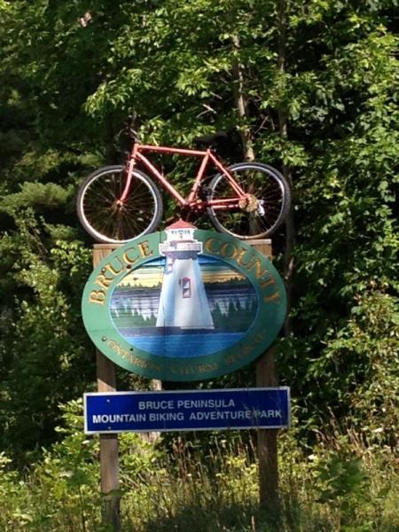 A colorful sign for Bruce County featuring an illustration of a lighthouse, with a pink bicycle mounted on top. The sign also includes a blue panel that reads "Bruce Peninsula Mountain Biking Adventure Park," surrounded by lush greenery. Bruce Peninsula Mountain Bike Adventure Park mountain bike trail.