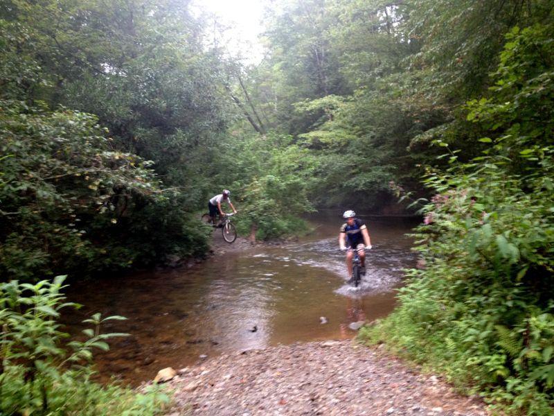 Two mountain bikers riding through a forested area near a shallow stream. One biker is jumping over the water while the other is splashing through it. Lush greenery surrounds the scene, creating a vibrant natural setting. Jake Mountain Trails mountain bike trail.