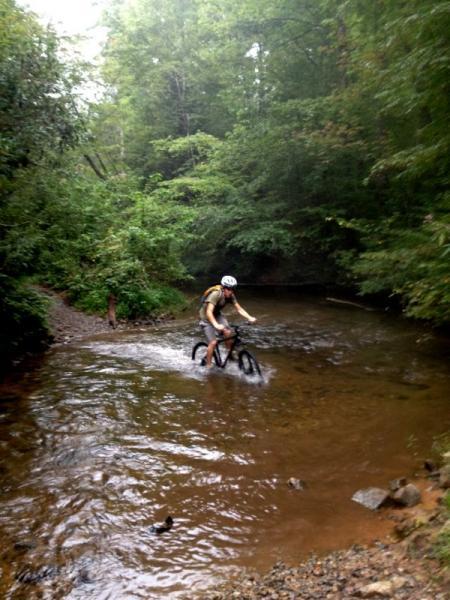 A mountain biker navigating through a shallow stream surrounded by dense greenery and trees. The rider is wearing a helmet and is partially submerged in water as they pedal their bike across the rocky terrain. Jake Mountain Trails mountain bike trail.