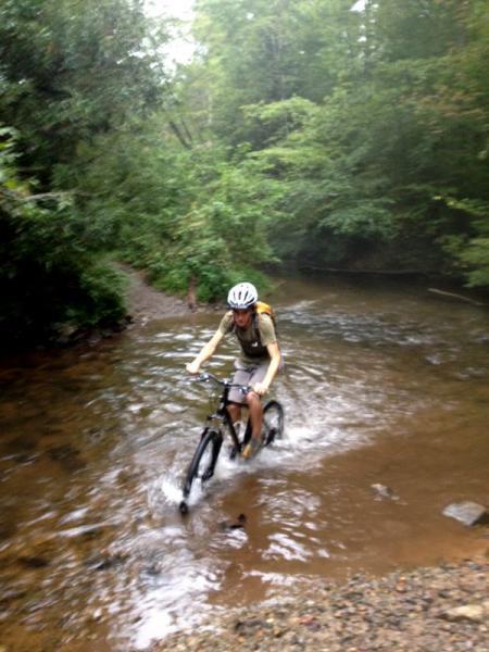 A person riding a mountain bike through a shallow, muddy stream in a wooded area, surrounded by lush green trees. The cyclist is wearing a helmet and is creating splashes as they navigate the water. The atmosphere appears misty, adding to the adventurous vibe of the scene. Jake Mountain Trails mountain bike trail.
