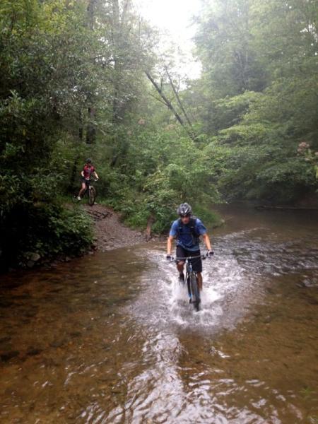 Two mountain bikers riding through a lush, green forest by a shallow stream. One rider splashes through the water in the foreground, while the other is positioned on a path to the left, surrounded by trees and vegetation. The scene is misty, creating a serene and adventurous atmosphere. Jake Mountain Trails mountain bike trail.