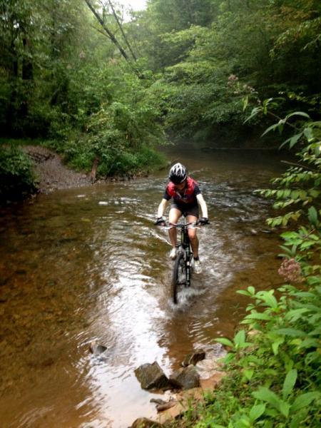 A person riding a mountain bike through a shallow creek, surrounded by lush greenery and trees. The cyclist is wearing a helmet and is partially submerged in the water as they navigate the rocky terrain. Jake Mountain Trails mountain bike trail.