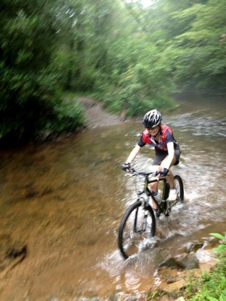 A person riding a mountain bike through a shallow river, surrounded by lush greenery. The cyclist is wearing a helmet and sportswear, splashing water as they navigate the rocky terrain. Jake Mountain Trails mountain bike trail.