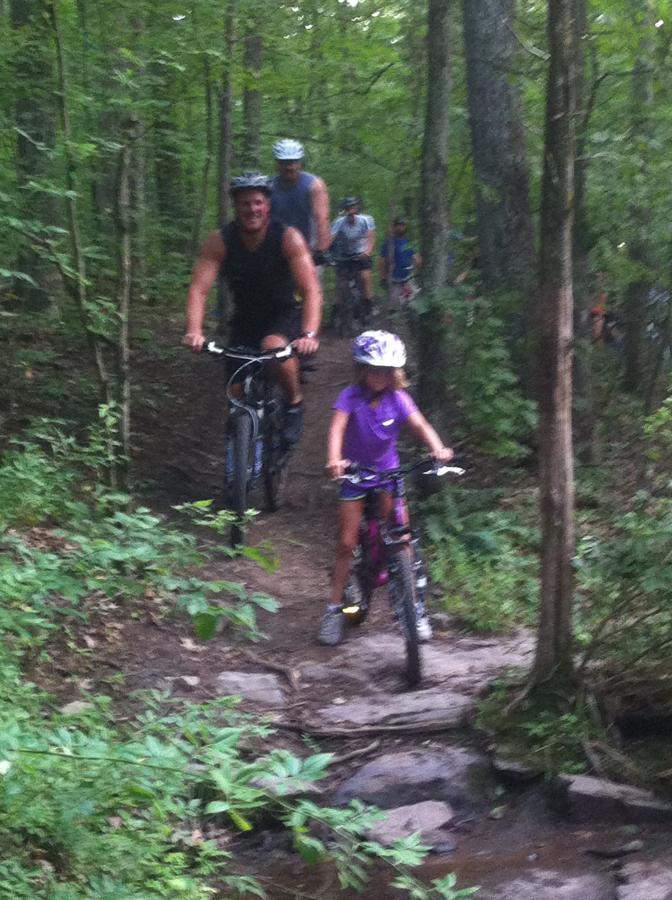 A group of people riding mountain bikes on a wooded trail, with a focus on a smiling adult cyclist in the foreground and a young girl in a purple shirt riding behind. The path is surrounded by lush green foliage, and there are rocks along the trail, suggesting a natural, rugged environment. Nockamixon State Park mountain bike trail.