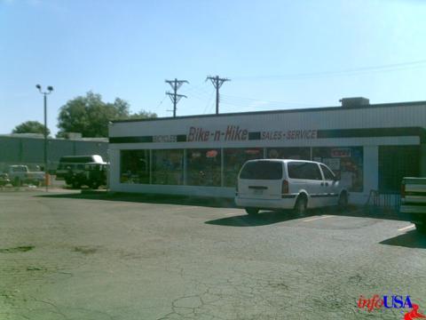 A storefront labeled "Bike-n-Hike Sales-Service," featuring large windows displaying bicycles and outdoor equipment. The building is situated in a paved area with parked vehicles and power lines in the background under a clear blue sky.
