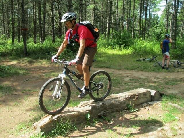 A person riding a mountain bike over a wooden log in a wooded area, surrounded by trees and grass. Another cyclist is in the background, adjusting their bike. The scene depicts an outdoor adventure and mountain biking activity. Kingdom Trails mountain bike trail.