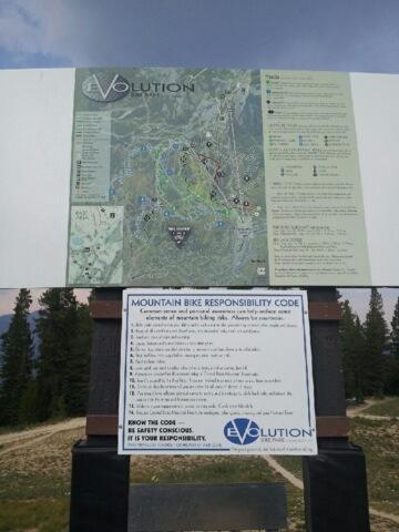 Map of the Evolution mountain biking area, featuring trails and a mountain bike responsibility code sign. The map includes various trails marked with symbols, and the sign outlines safety and responsibility guidelines for riders. The background shows trees and a cloudy sky. Evolution Bike Park at Crested Butte Mountain Resort mountain bike trail.
