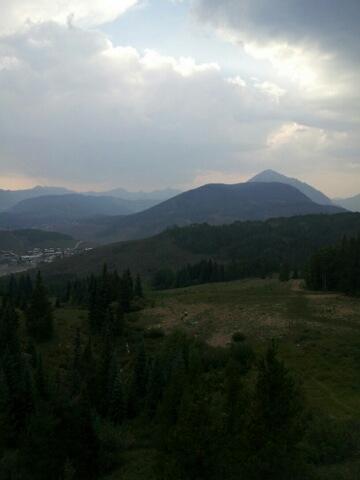 A scenic view of rolling mountains under a cloudy sky, with patches of greenery in the foreground and varying shades of blue in the distant peaks. Evolution Bike Park at Crested Butte Mountain Resort mountain bike trail.