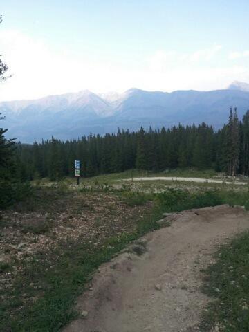 A scenic view of a mountainous landscape with lush green trees in the foreground and distant peaks under a blue sky. A dirt path leads through the area, and a sign can be seen in the middle distance, indicating a recreational area. Evolution Bike Park at Crested Butte Mountain Resort mountain bike trail.