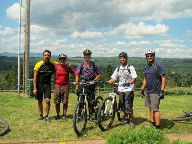 Five men stand on a grassy area with mountain bikes beside them, wearing helmets and cycling gear. They are smiling and posing for the camera, with a scenic landscape of hills and a partly cloudy sky in the background. Kingdom Trails mountain bike trail.