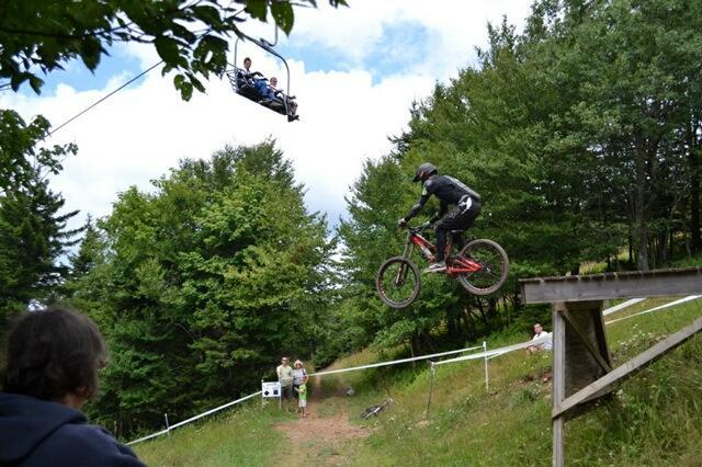 A mountain biker in a black outfit performs a jump off a wooden ramp while spectators watch nearby. A chairlift carrying passengers is visible above, with trees and a cloudy sky in the background. Snowshoe Bike Park mountain bike trail.