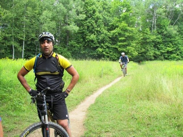 A person wearing a black and yellow cycling jersey stands next to a mountain bike on a dirt path surrounded by tall grass and trees. In the background, another cyclist is riding down the trail. The scene depicts a lush green outdoor setting, ideal for mountain biking. Kingdom Trails mountain bike trail.