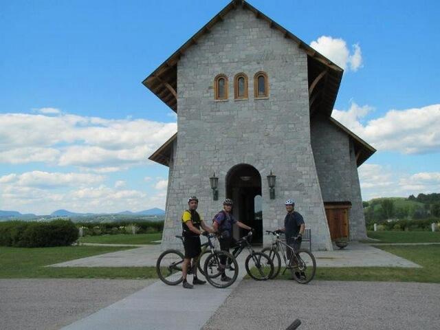 Three cyclists stand in front of a gray stone building with large windows and a wooden door, set against a backdrop of blue sky and distant mountains. The men, wearing cycling helmets and gear, are positioned with their mountain bikes on a path leading to the entrance of the structure. Lush green grass surrounds the area. Kingdom Trails mountain bike trail.