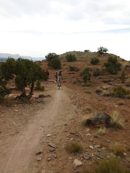 Two mountain bikers are riding along a dirt trail through a rocky desert landscape. The trail winds up towards a small hill covered with sparse vegetation and a few scattered bushes. The sky is overcast, hinting at possible rain. Lunch Loops mountain bike trail.