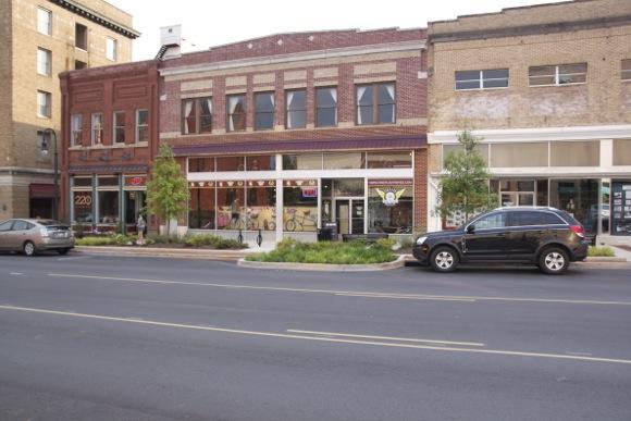 A street view of a two-story brick building featuring storefronts on the ground level. The building has large windows adorned with colorful displays. In front, there are landscaped areas with small trees and grass. Several cars are parked along the road and the scene captures a mix of historic and modern architectural styles.