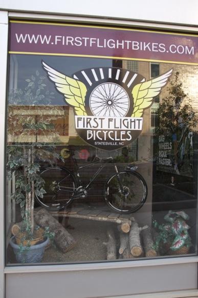 Alt text: A storefront window displaying the logo of First Flight Bicycles, featuring a bicycle and decorative logs and plants. The logo includes wings and text that reads "First Flight Bicycles" and "Statesville, NC," along with the website URL at the top.