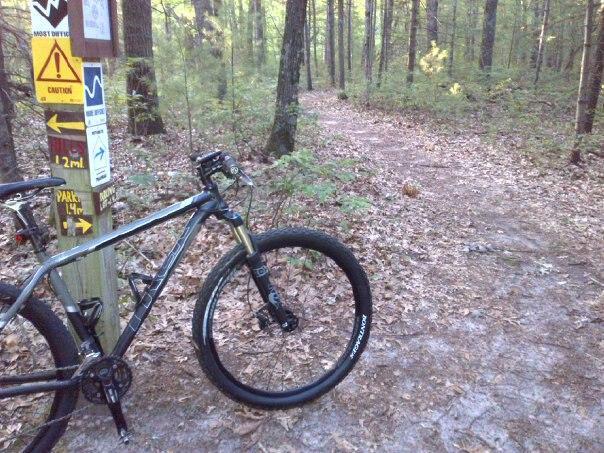 A mountain bike leaning against a signpost in a wooded area, with trail markers and directional arrows visible. The path ahead is partially visible, surrounded by trees and fallen leaves, indicating a natural trail for biking or hiking. Pentwater Pathways mountain bike trail.