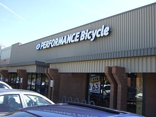 Exterior view of a Performance Bicycle store showing the sign above the entrance and parked cars in the foreground. The building features a tan façade with multiple access points.