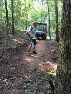 A person wearing a backpack walks along a dirt trail in a wooded area, with a utility vehicle parked in the background. Sunlight filters through the trees, creating a natural, outdoor setting. Paynes Creek mountain bike trail.