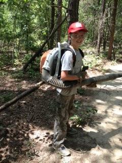 A young person wearing a red cap and camouflage pants stands in a wooded area, holding a backpack leaf blower. They are smiling and appear to be ready to work outdoors. The background features trees and underbrush typical of a forested environment. Paynes Creek mountain bike trail.