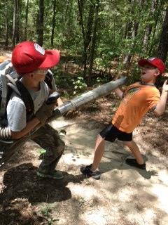 Two children are playfully pretending to use a leaf blower in a wooded area. One child, wearing a backpack and holding the blower, is aiming it toward the other child, who is leaning back with an exaggerated expression of surprise. Both are wearing baseball caps, and the ground is covered in dirt and leaves. Paynes Creek mountain bike trail.