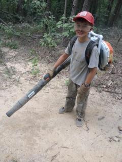 A young boy wearing a red cap and a gray t-shirt stands on a sandy area in the woods, holding a backpack leaf blower. His clothing and hands appear slightly dirty, indicating he is engaged in outdoor work. Trees and greenery are visible in the background. Paynes Creek mountain bike trail.