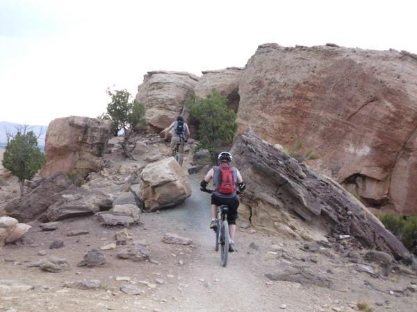 Two mountain bikers navigating a rocky trail through a rugged landscape, with large boulders and sparse vegetation surrounding them. The sky is overcast, indicating a cloudy day. Lunch Loops mountain bike trail.